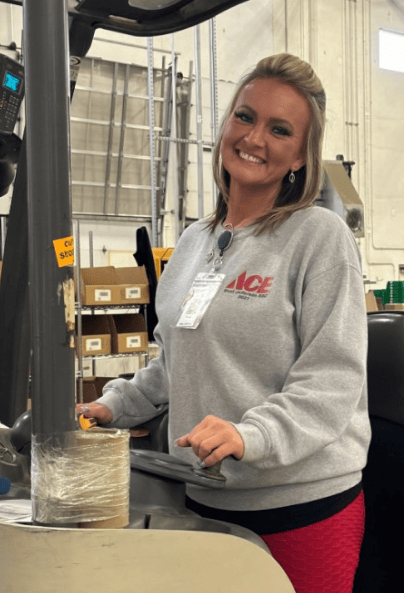 A woman wearing a gray ACE sweatshirt and name badge smiles while standing next to a forklift in a warehouse setting. Shelves with boxes and supplies are visible in the background.
